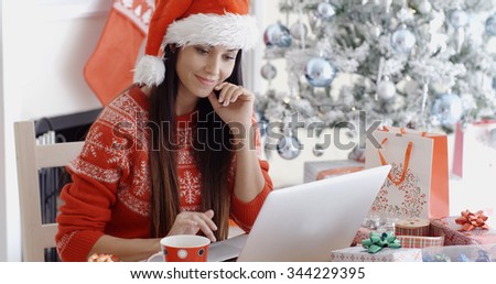 Smiling young woman surfing the internet on her laptop computer for Christmas bargains as she sits at a table in front of the decorated tree in her red Santa hat.