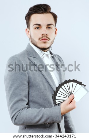 Attractive young man, with beard and mustaches, wearing in white shirt and scissors-bow, posing with cards in his hand, on white background, in studio, waist up