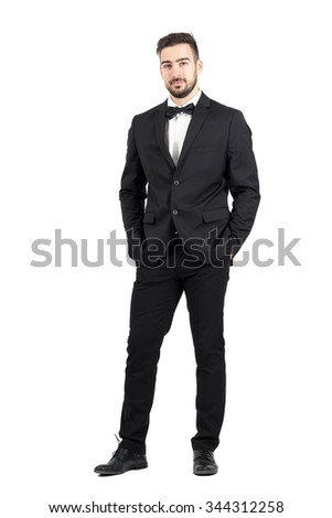 Wealthy confident relaxed young man in tuxedo looking at camera with hands in pockets. Full body length portrait isolated over white studio background. Royalty-Free Stock Photo #344312258
