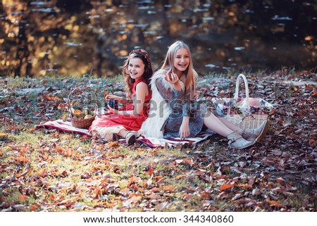 Two pretty laughing blonde and brunette girls in beautiful gowns with fruit in their hands and picnic baskets sitting on plaid on lake shore in autumn park, horizontal picture 