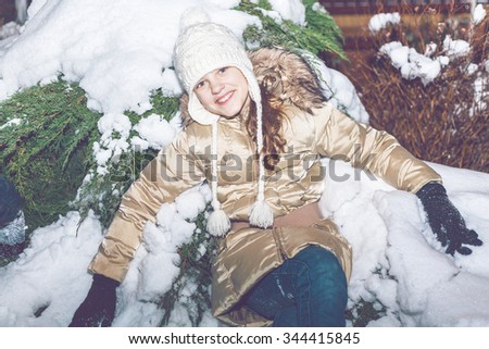cheerful young girl in a woolen hat and yellow jacket sitting in snow in winter evening forest