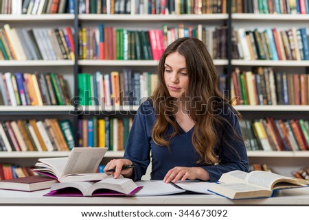 serious young girl with loose long dark hair  sitting at a desk in the library with an open note book writing out information from books, looking at notes, a concept of studying.