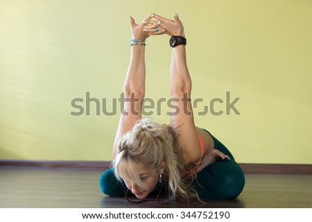 Sporty beautiful blond young woman in sportswear working out indoors, doing variation of Padmasana, sitting cross-legged, bending forward in Lotus Posture, exercise for shoulders flexibility