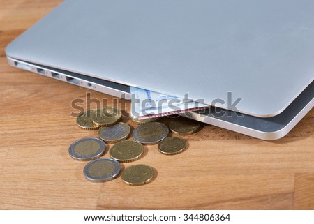 An image of a laptop including European cash and some coins on a wooden table. 