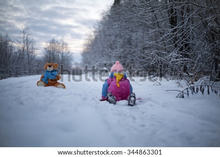 Baby girl is playing outdoor. She is crawling to teddy bear sitting on the snow.