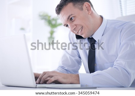 Profile shot of a  businessman working on his laptop in the office.Businessman talking on the phone.He is wearing a blue shirt and a black tie.