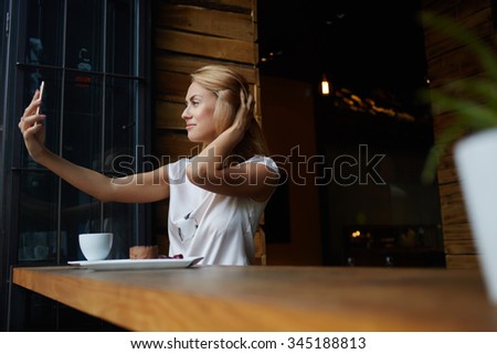 Young gorgeous woman making self portrait with mobile phone camera while sitting alone in modern coffee shop, attractive European female posing while photographing herself for social network picture 