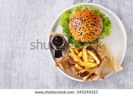 Fresh homemade burger with black sesame seeds in white plate with french fries potatoes, served with ketchup sauce in glass jar over gray wooden surface. Top view Royalty-Free Stock Photo #345244325