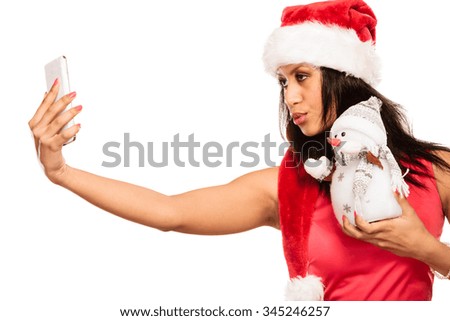 Winter and Christmas time concept. Woman in santa helper hat, mixed race girl in red dress holding happy nice snowman toy, taking self picture selfie with smartphone camera. Studio shot on white