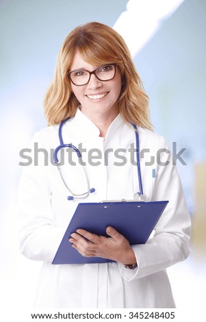 Portrait of confident female doctor holding in her hand a clipboard and smiling to the camera while standing at hospital.