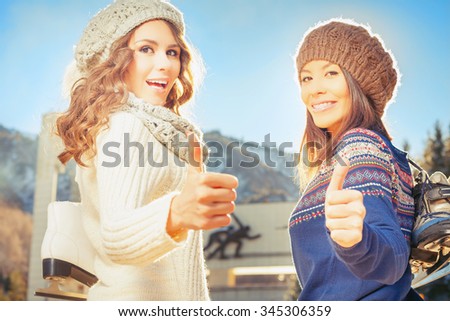 Happy multiracial women going to ice skating outdoor with thumbs up gesture. Holding skates shoes. Healthy lifestyle and winter sport concept at sports stadium, mountain landscape
