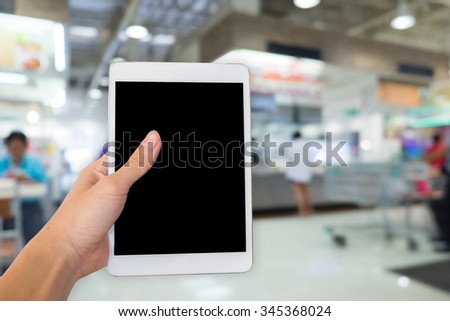 Hands are holding touch screen White Tablet Computer on blurred shopping super market background.
