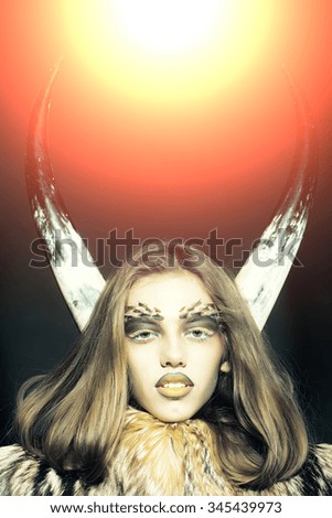 Closeup portrait of one beautiful wild young woman with bright golden animal monkey makeup with thorns on face and antlers in fur coat on black background with light splash, vertical picture