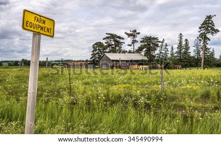 A farm equipment sign at the edge of a road warning traffic of slow moving vehicles. A farm field and buildings are in the background.