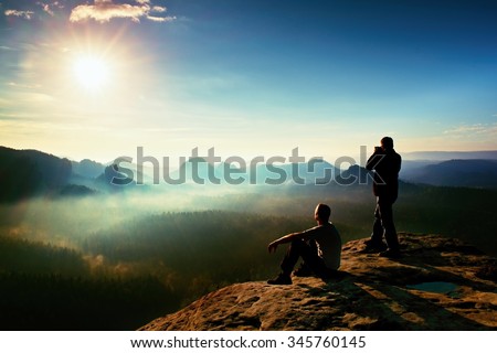Hiker and photo enthusiast stay with tripod on cliff and thinking. Dreamy fogy landscape, blue misty sunrise in a beautiful valley below, soft focus