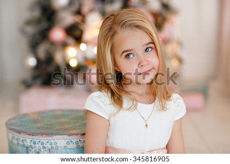 Very nice charming little girl blonde in white dress lying on the bed and looks sad in the picture on the background of Christmas trees in bright interior of the house