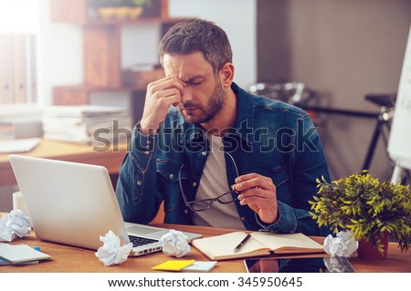 Feeling sick and tired. Frustrated young man massaging his nose and keeping eyes closed while sitting at his working place in office Royalty-Free Stock Photo #345950645