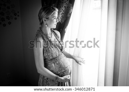 Pregnant woman is looking from the window in her flat. Black and white photography.