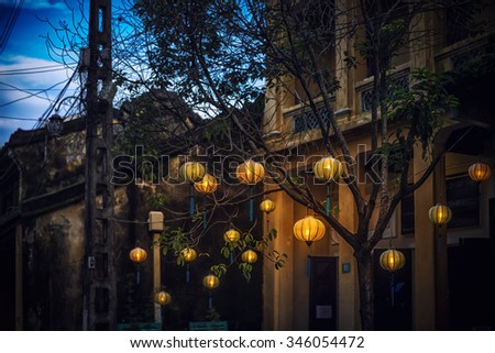 Lanterns  hanging on tree in Hoi An. Traditional asian lamps made of paper. Yellow wall of old building with two small windows. Equipment for cultural events in Vietnam, Asia.