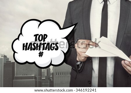 Top hashtags text on speech bubble with businessman holding paper plane in hand on city background