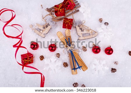 Xmas or new year composition with holiday decorations - little cristmas baubles, red satin ribbon on snow with toy skates, skis and gifts on the sledge. Top view. Christmas card