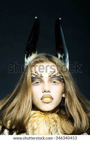 Closeup portrait of one beautiful wild young woman with bright golden animal monkey makeup with thorns on face and antlers in fur coat on black background, vertical picture