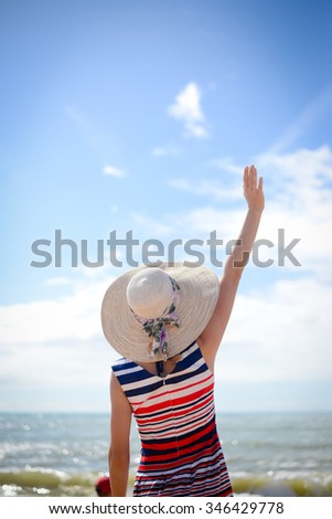 Back view of elegant lady in straw hat standing on seaside and waving her hand on picturesque sky and sea background 