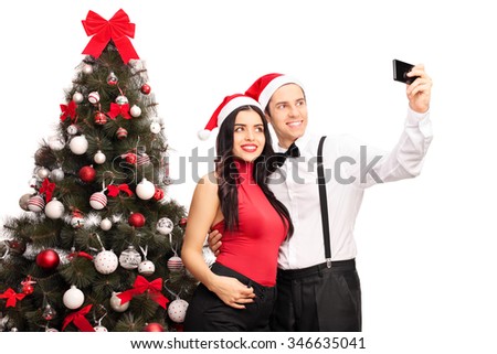 Young man and woman with Santa hats taking a selfie in front of a Christmas tree isolated on white background