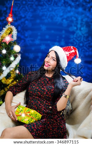 Picture of beautiful young lady in red Santa hat sitting with present box. Sparkling image of pretty girl happy smiling beside Christmas tree on blue digital background with snowflakes.