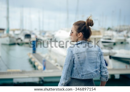 Picture of beautiful young woman standing on city embankment. Side view of pretty girl in sunglasses waiting on blurred marine outdoor background.
