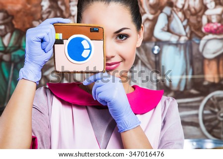 Beautiful woman, wearing in pink dress, apron and violet gloves, holding gingerbread which looks like camera in front of her face, in the kitchen with painted walls, close up