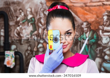 Attractive young woman, wearing in pink dress, apron and violet gloves, with ribbon in her hair, holding gingerbread which looks like camera in front of her face, in the kitchen with painted walls