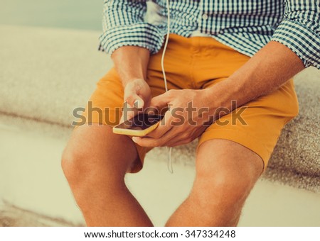 young hipster guy holding a smartphone responds to mail texting and send pictures while sitting on the pier,Young man wearing white blank t-shirt and blue jeans, standing on the street,