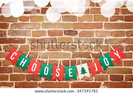 Red, green and white CHOOSE JOY and twig tree paper card DIY flag garland decoration top blurred lights border on rustic brick wall background. Horizontal postcard with space for text, or lettering.