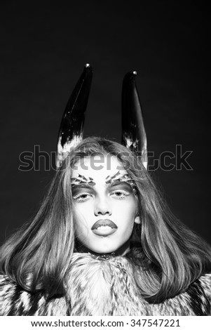 Closeup portrait of one beautiful wild young woman with bright animal monkey makeup with thorns on face and antlers in fur coat on black background black and white, vertical picture