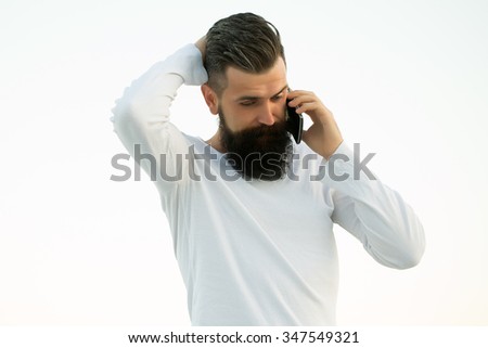 One handsome young bearded thoughtful business man in white sweater holding and speaking on cell phone outdoor on light sky natural background, horizontal picture