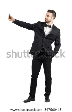 Young man in suit with bow tie taking selfie with cellphone. Full body length portrait isolated over white studio background. 