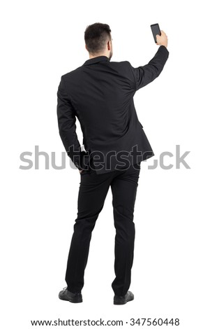Rear view of young executive in black suit taking photo with his cellphone.  Full body length portrait isolated over white studio background. 