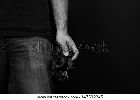 Black and white close up image of a man wearing jeans and a t shirt holding a film sir camera at his side