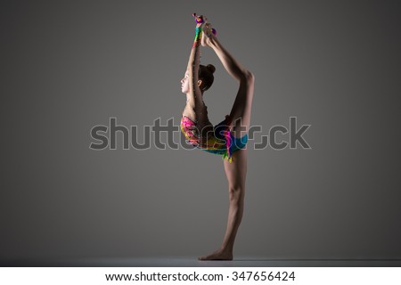 Beautiful cool young fit gymnast woman in colorful sportswear working out, standing on one leg, performing back scale, rhythmic gymnastics backbend element with maces, studio, dark background