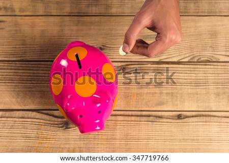 Hands close up putting a coin into a pink pig on a wooden table