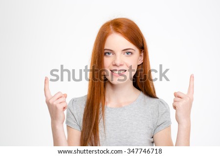 Happy cheerful redhead young woman with long hair pointing up with fingers of both hands over white background