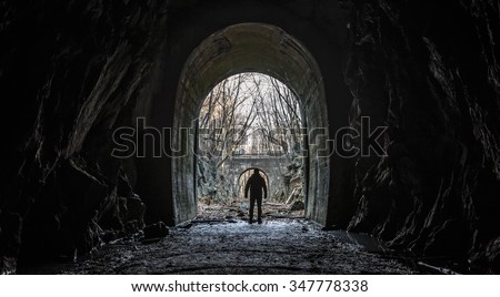 Wide silhouette view of a lone survivor emerging from entrance or exit of an abandoned cave tunnel with an apocalyptic feel and dark cinematic grunge effect.