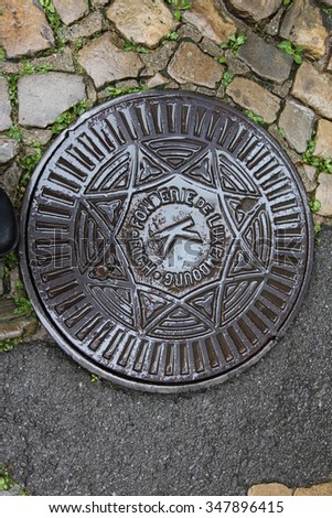 The old sewage manhole is on the dark cobblestone pavement in Luxembourg, after the rain.