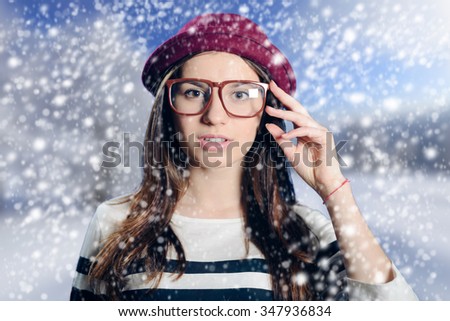 Portrait of young female in fancy hipster glasses wearing striped sweater and dark red felt hat in snowy weather background