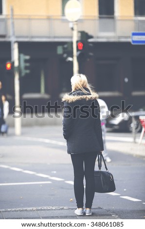 A blonde girl is standing in the streets when traffic lights is red. She is wearing black clothes and white shoes and has a black bag with her. Image has a vintage effect applied.