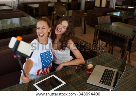 Cheerful hipster girls posing while photographing herself for social network picture during lunch break in cafe, happy women making photo with smart phone camera via self stick while sitting in bar 