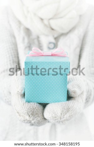 Hands in white mittens holding small blue gift box with pink ribbon. 