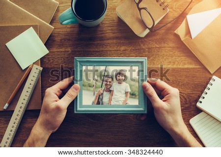 My son is my inspiration. Close-up top view of man holding photograph of himself and his son fishing over wooden desk with different chancellery stuff laying around