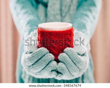 Woman holding winter cup close up on light background. Woman hands in teal gloves holding a cozy mug with hot cocoa, tea or coffee and a candy cane. Winter and Christmas time concept.
